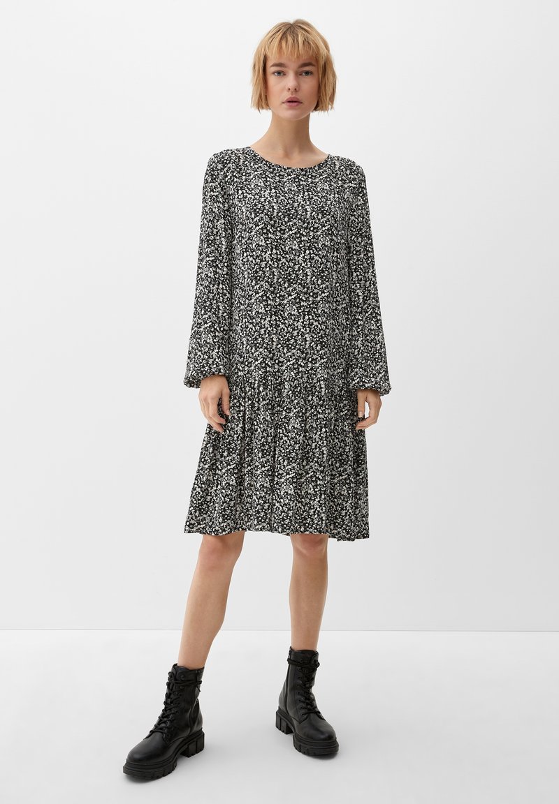 Schwarzes und weißes Blumenkleid mit langen Ärmeln, gerafftem Bund und gestuftem Rock. Getragen mit schwarzen Stiefeln im Combat-Stil. Glatte Stoffstruktur.