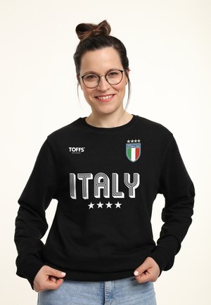 Smilende kvinde med briller iført en sort sweatshirt med teksten Italien, fire stjerner og et Italia-skjoldlogo, der holder i trøjekanten.