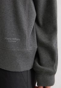 Felpa grigia con polsini e orlo a coste, dal texture liscia e con un logo discreto in basso. Primo piano del dettaglio della manica.