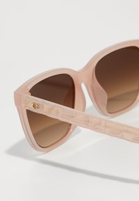 Óculos de sol de cor rosa claro com uma armação grande e angular e lentes castanhas em gradiente. As hastes transparentes têm uma textura linear e um detalhe em dourado.