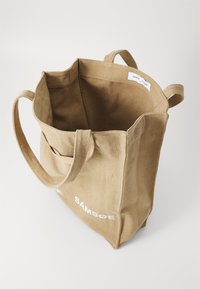 Beige Canvas-Tragetasche mit zwei langen Tragen, offenem Oberteil und sichtbarem Etikett. Sie zeichnet sich durch ein einfaches Design aus und der Markenname ist in Weiß gedruckt.