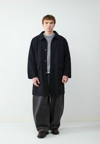 Schwarzer Wollmantel mit Kragen, vorderen Knöpfen und seitlichen Taschen, kombiniert mit einem hellgrauen Pullover und übergroßen dunklen Jeans.