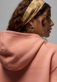 Hoodie rose pastel avec un cordon de serrage, texture douce et style sweatshirt. Bandeau brun à motifs et boucles d'oreilles en or en guise d'accessoires.