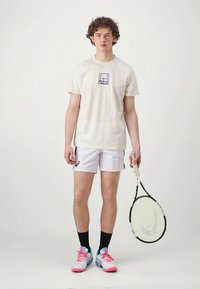 Wit katoenen T-shirt met een grafisch logo, gecombineerd met witte shorts en felgekleurde sneakers. Houdt een tennisracket vast.