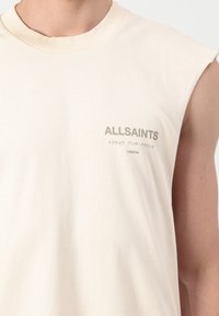 Bezrukávové krémové tričko s nápisom "ALLSAINTS," japonskými znakmi a "LONDON" potlačenými na ľavej strane hrudníka.