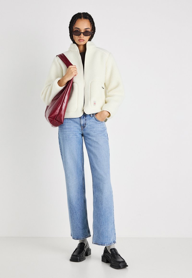 Kvinde iført hvid fleecejakke, lyseblå jeans, sorte loafers, solbriller, mens hun holder en mørkerød læder skuldertaske, stående mod en hvid baggrund.