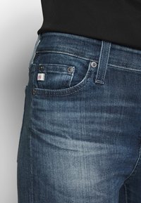 Tmavě modré džíny vyrobené z denimu s mírně vyblednutou texturou. Mají dvě přední kapsy, stříbrné kovové doplňky a malou štítkovou nášivku.