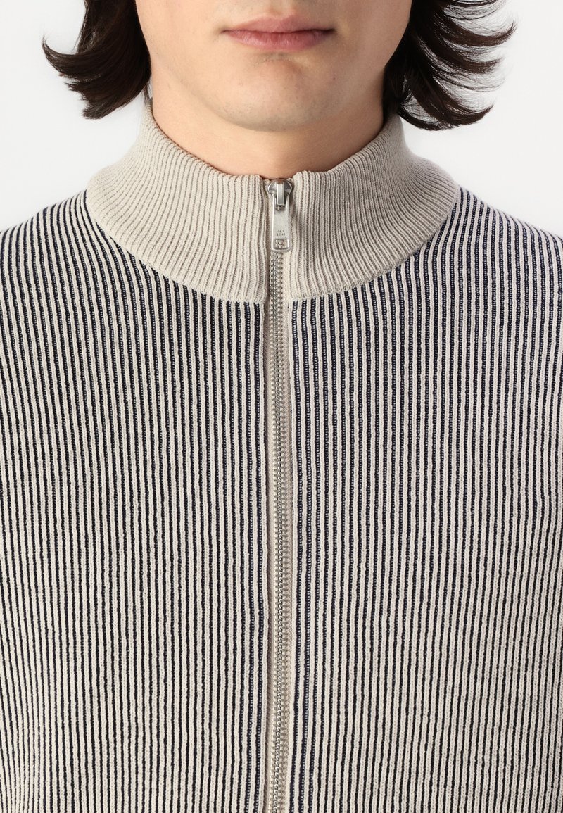 Maglione zip-up lavorato a maglia a righe crema e navy, con colletto alto e zip in evidenza. Texture morbida e a coste in tutto il capo.