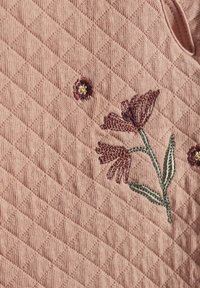 Vadderat rosa tyg med blommig broderi i djup röd och grön, med ett diamantmönster och inslag av små broderade blommor.