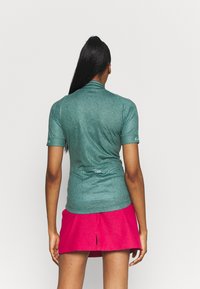 Grön kortärmad polo med ett strukturerat vågigt mönster och krage, ihop med en rosa utsvängd kjol. Tydlig logotypdetalj på ärmen.