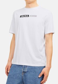 Hvid kortærmet T-shirt lavet af bomuld, med et sort grafisk logo "JACK&JONES" tværs over brystet. Klassisk rund halsudskæring.