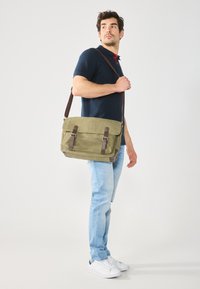 Olive green messenger bag made of textured fabric with brown leather accents, featuring adjustable strap and two front buckles.