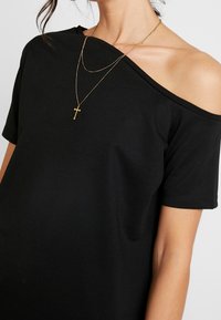 Černé tričko s odhalenými rameny vyrobené z měkké látky, s krátkými rukávy a volným střihem, doplněné o vrstvené zlaté řetízky.