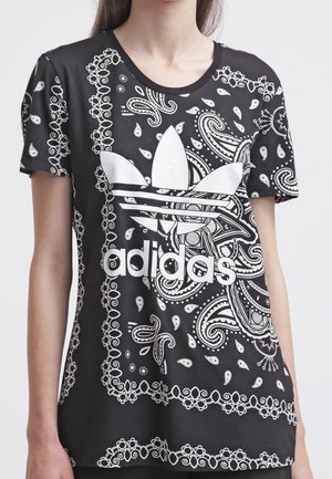 Osoba nosząca czarną koszulkę Adidas z białymi wzorami paisley i bandana oraz dużym białym logo Adidas trefoil na przodzie.