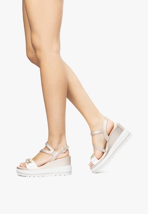Gambe di donna che indossano sandali con zeppa platform beige e bianchi con cinturini alla caviglia, che camminano su uno sfondo bianco senza decorazioni.