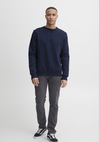 Navy blue crew neck sweatshirt with ribbed cuffs, paired with gray jeans and black sneakers featuring white accents. Simple, casual design.