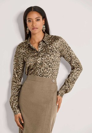 Vrouw met lang donker haar draagt een blouse met luipaardprint en een beige rok met hoge taille, staand met één hand op de heup tegen een witte achtergrond.