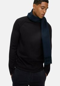 Mann in einem schwarzen Langarmshirt und einem dunkelblauen Schal, der um seinen Hals drapiert ist, mit den Händen in den Taschen und nach unten zur Seite blickend.