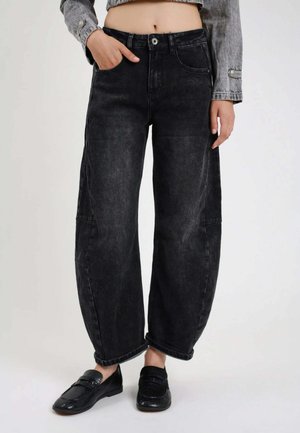 Persona che indossa jeans neri larghi a vita alta con le mani nelle tasche e mocassini neri, top grigio a maniche lunghe corto che mostra l'addome.