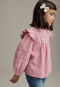 Rosa bluse med flagrende ærmer og strukturerede blomsterdesigns. Har pintuck-detaljer og elastiske manchetter. Bæres med denim.