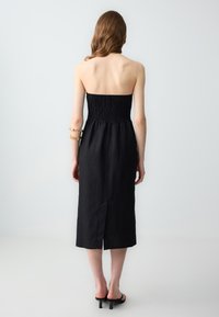 Schwarzes, trägerloses Kleid mit gerüschtem Oberteil und Midi-Länge. Der Stoff wirkt leicht und hat eine feine Textur. Es verfügt über einen Schlitz in der Mitte des Rückens.