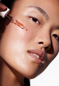 Un contagocce trasparente contiene un siero ambrato chiaro che si applica su una pelle liscia e luminosa con una leggera texture, circondata da uno sfondo sfocato.