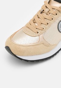 Sneaker beige chiaro con accenti metallici dorati, caratterizzati da dettagli perforati, design con lacci e una suola in gomma nera testurizzata.
