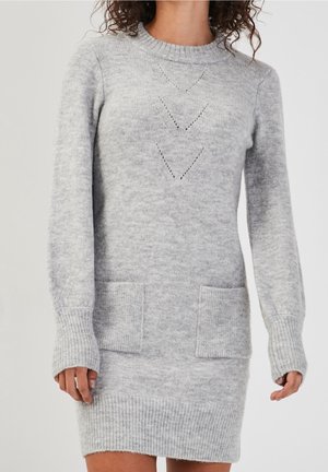 Woman wearing a light gray knit sweater dress with long sleeves, two front pockets, and chevron knit patterns on the chest.