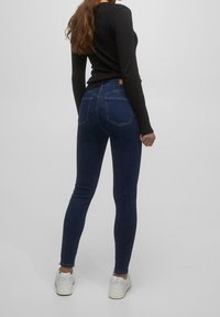 Jeans ajustados de denim azul oscuro con una textura suave y dos bolsillos traseros, combinados con una blusa negra de manga larga ajustada y zapatillas blancas.