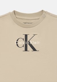 Béžové tričko Calvin Klein s vroubkovaným límcem, patentním knoflíkem na rameni a černým logem "cK" s bílým textem "Calvin Klein" uprostřed.