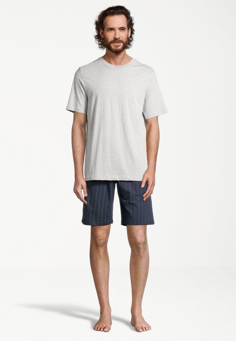 Homme aux cheveux bouclés et barbe, debout pieds nus, portant un t-shirt à manches courtes gris clair et un short bleu marine à fines rayures sur fond blanc.