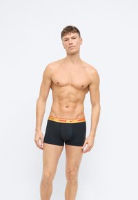 Čierne boxerky Nike s červenožltým pásom, s priliehavým strihom a hladkým materiálom, vystavené na hrudníku muža bez trička.