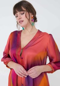 Satijnen jurk met een diepe V-hals en kleurrijke gradient, met tinten roze, oranje en paars; heeft lange mouwen en decoratieve knopen.