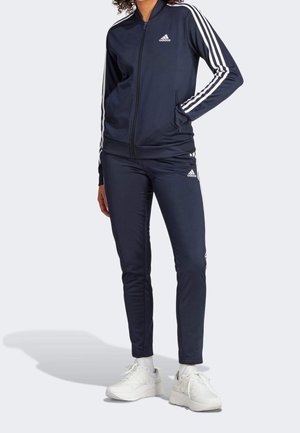 Osoba w granatowym dresie Adidas z białymi paskami na rękawach i spodniach, w białych trampkach, stojąca z jedną ręką w kieszeni.