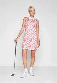 Hihaton golfmekko, jossa on vaaleanpunainen, violetti ja valkoinen abstrakti kuvio, kaulukset ja vartalonmyötäinen leikkaus, yhdistettynä valkoisiin golfkenkiin.