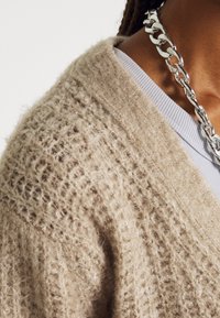 Beige knitted cardigan with a textured pattern, made of soft yarn, worn over a light gray top, featuring a shiny silver chain necklace.