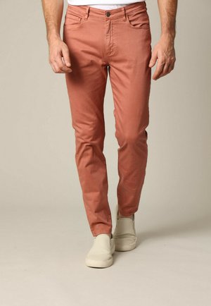 Pantalons en coton rouge-orange avec une coupe slim, design à cinq poches et texture subtile, associés à des chaussures slip-on blanches.