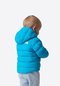 Väikelaps erksassinises kapuutsiga talvejope ja heledates pükstes, külje pealt alla vaatamas, lihtsal heledal taustal.