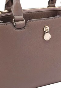 Bruine leren handtas met een gestructureerd oppervlak, voorzien van gouden hardware en een ronde hanger. Ritssluiting en dubbele handgrepen voor het dragen.