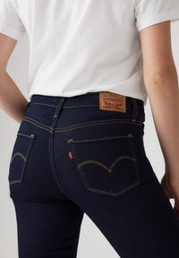 Vaqueros de mezclilla azul oscuro con un diseño ajustado, costuras amarillas en contraste y un parche de cuero marrón con el logo de Levi's en la cintura.
