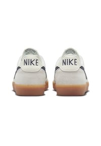 Sneakers de couro branco com detalhes em azul marinho, sola de borracha de cor caramelo e uma marca "NIKE" proeminente no calcanhar. Detalhes em camurça na parte de trás.