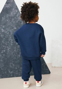 Un enfant aux cheveux bouclés se tient de dos, vêtu d'un sweat-shirt bleu marine et d'un pantalon, avec des baskets blanches dotées de tirettes rouges.
