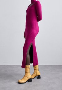 Robe en maille côtelée magenta ajustée avec un col roulé et des fentes latérales. Associée à des bottines jaunes à talon épais avec des détails de couture.