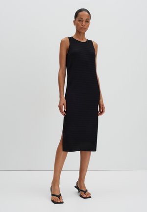 Schwarzes ärmelloses Kleid mit einem strukturierten Muster, knielang, mit Seitenschlitzen und rundem Ausschnitt. Trägt schwarze Flip-Flop-Sandalen.