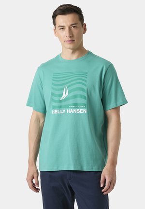 Uomo che indossa una maglietta teal Helly Hansen con grafica di una barca a vela e coordinate, abbinata a pantaloni blu scuro, in piedi contro uno sfondo chiaro.