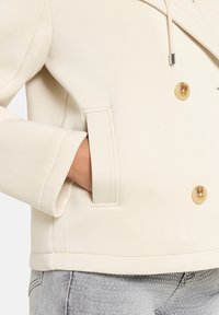 Cremefarbener zweireihiger Blazer mit großen, hellbraunen Knöpfen, einem strukturierten Kragen und Seitentaschen, aus einem glatten Stoff gefertigt.