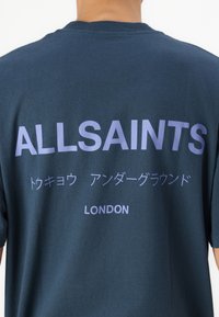 Tamsiai mėlynas medvilnės marškinėliai trumpomis rankovėmis su baltais ir šviesiai violetiniais užrašais. Užrašai apima "ALLSAINTS", japoniškus simbolius ir "LONDON".