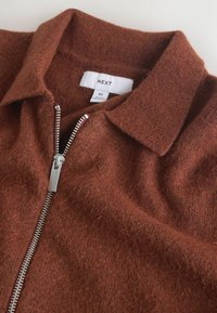 Rust-colored zip-up shirt made of textured fabric. Features a standard collar and a silver metal zipper along the front. Brand label visible.