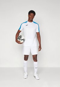 Biały strój sportowy z niebieskimi akcentami, krótki rękaw i dopasowane spodenki. Model trzyma czarno-białą pi�łkę nożną i ma na sobie białe skarpetki oraz buty.