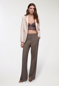 Blazer beige, pantaloni marroni su misura e un bralette in pizzo viola. Tessuti lisci, design aderente e tacchi a punta marroni con accenti.